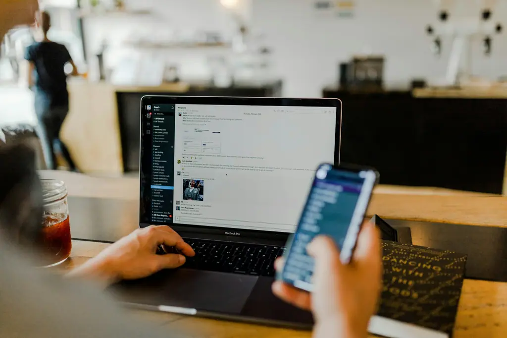 A person browsing on a smartphone while working on a laptop in a café